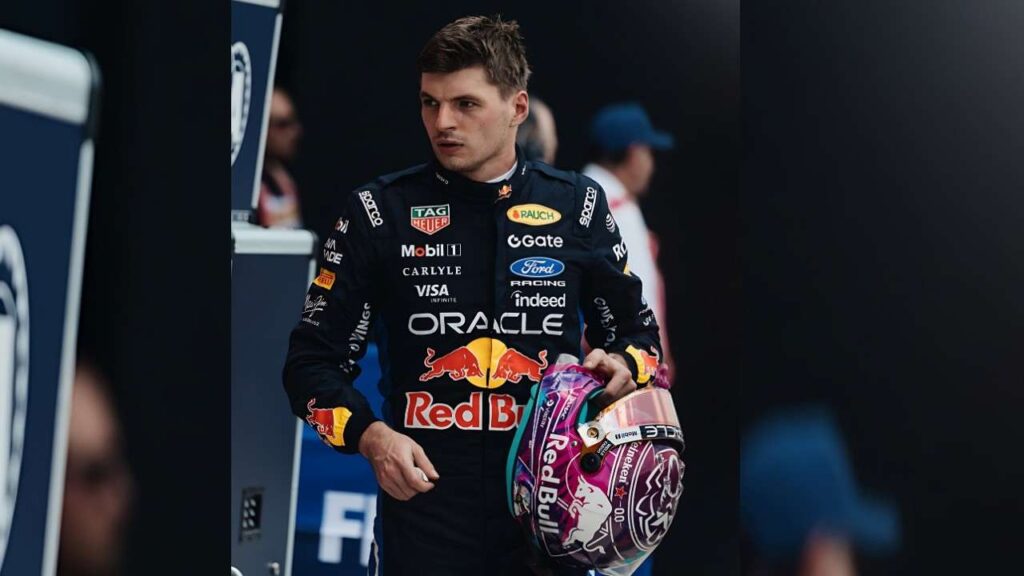 Max Verstappen F1 driver in a dark blue Red Bull suit stands in a dim paddock, holding a vibrant pink-purple helmet with Red Bull branding.