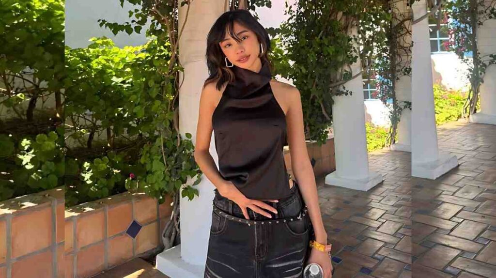 Maymay Entrata with short dark hair in a black satin halter top and black jeans with a studded belt poses on a sunlit patio beside white columns, holding a bottle.