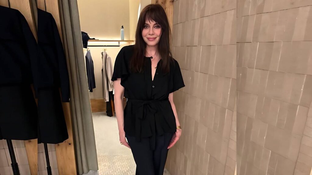 Melinda Clarke with dark hair in a black belted blouse and dark pants stands in a boutique, beige tiled wall and racks behind.