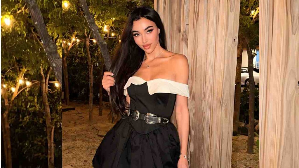 Metisha Schaefer in an off-shoulder black and white dress with a belt, posing beside wooden posts under warm string lights.