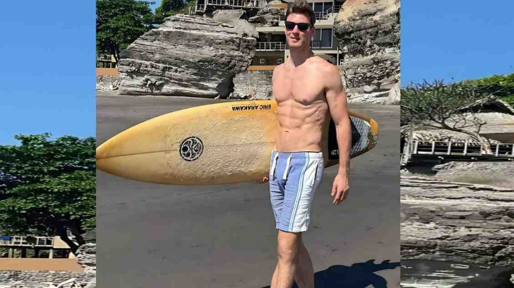 Michael Jeffrey in blue-and-white striped trunks, wearing sunglasses, stands on a sunny beach with a yellow surfboard.
