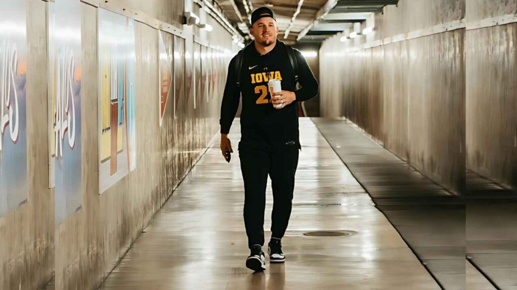 Mike Trout in a black Iowa jersey and cap walks through a dim, hallway-like tunnel, coffee cup in hand and backpack slung.