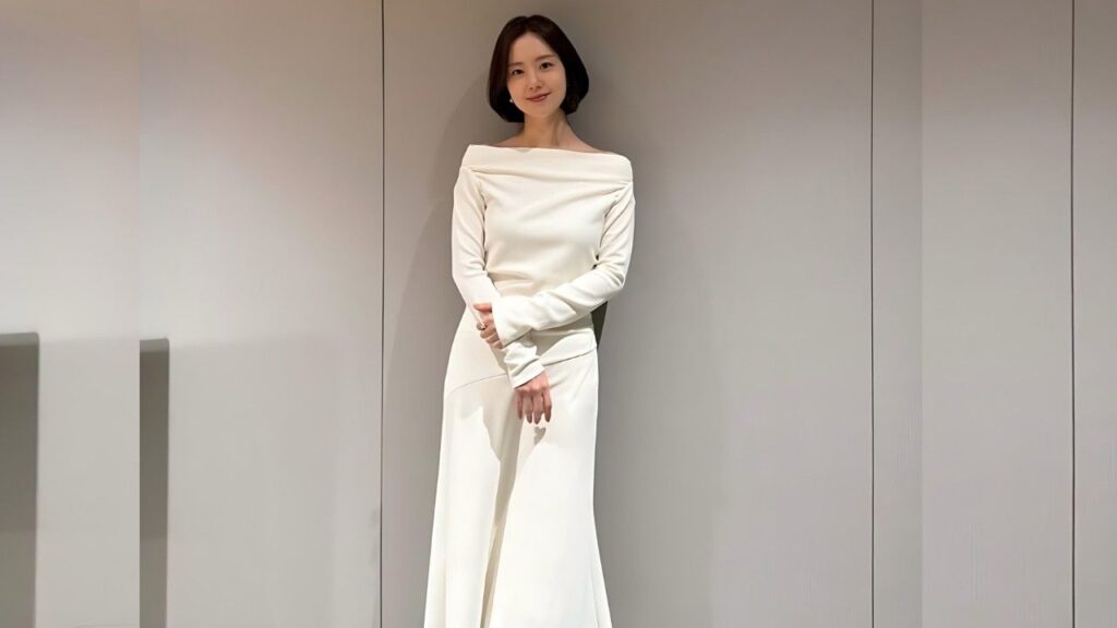 Moon Chae-won with a short dark bob stands against a pale paneled wall, wearing an off-shoulder white top and long white skirt, arms crossed, smiling.