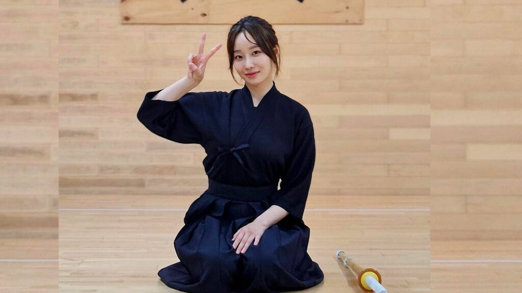 Moon Ye-won kneels on a wooden gym floor in a navy kimono, smiling and flashing a peace sign; a practice sword lies nearby.
