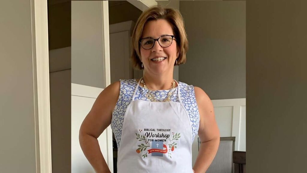Nancy Guthrie with short hair and glasses wears white apron over blue top; apron reads Biblical Theology Workshop for Women.