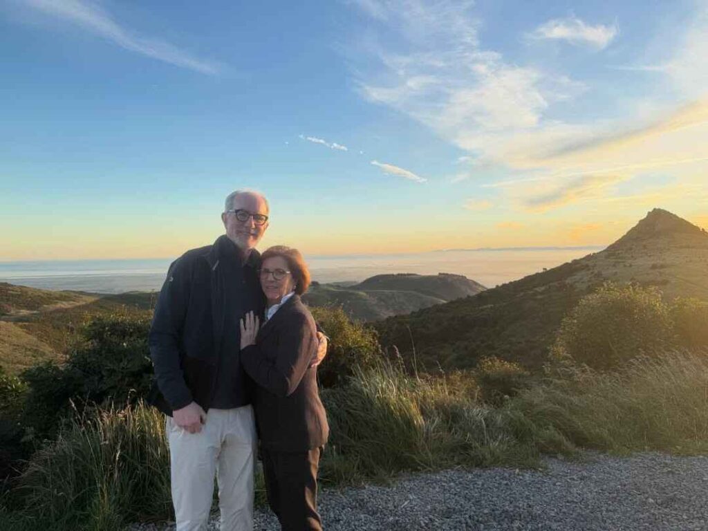 Nancy Guthrie Husband and Nancy Guthrie in glasses share a hug on a gravel overlook at sunset, with rolling hills, sea horizon, and a pointed hill to the right.