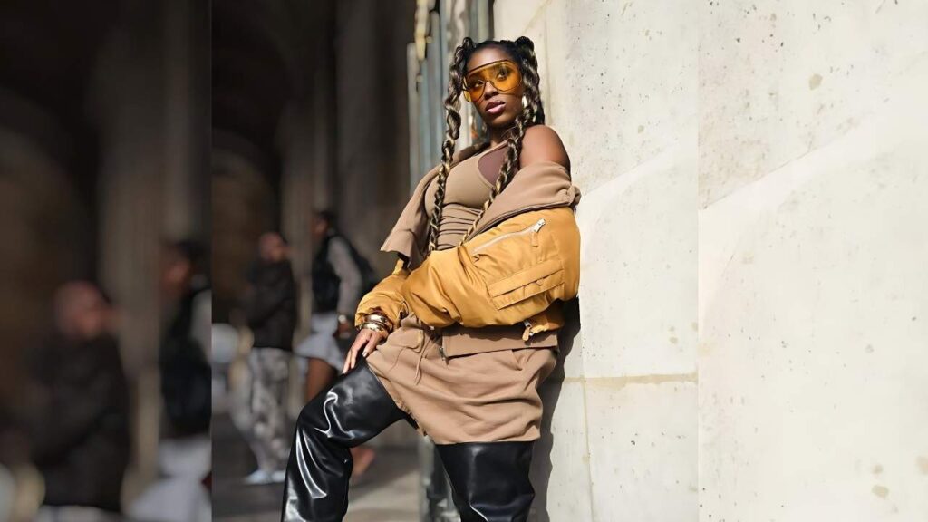 Naomi a pale stone wall in a tan outfit with a mustard jacket, beige top, black pants, and orange sunglasses.