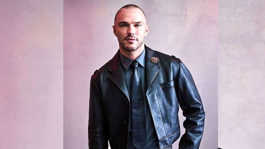 Nicholas Hoult in a dark leather blazer, blue shirt and tie, with a flower lapel pin, stands against a soft pink backdrop.