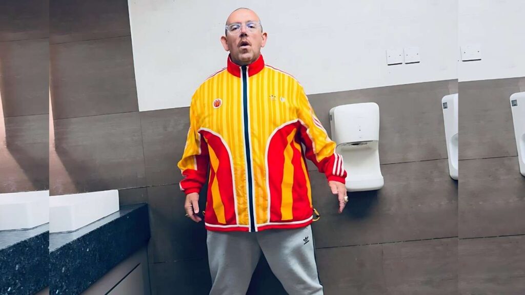 Nick Frost in bright yellow and red striped jacket, gray sweatpants, bald with glasses, stands in a modern bathroom, mouth open.