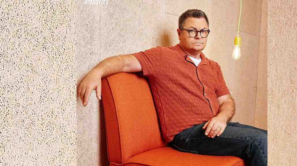 Nick Offerman with glasses sits on orange couch against a beige wall, wearing a red-orange shirt; a hanging bulb glows nearby.