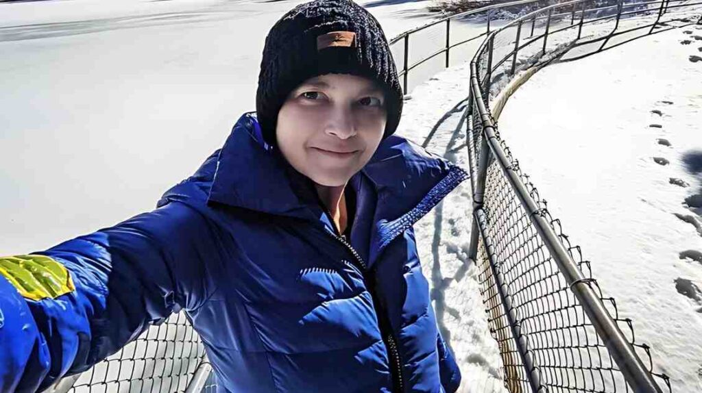 Nicki Aycox in a blue puffer jacket and black beanie selfies on a snowy, fenced path beside a frozen lake, footprints.