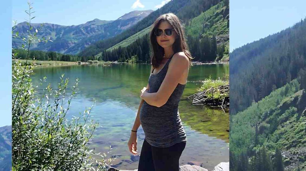 Nicole Linkletter in gray tank top and black leggings by a mountain lake, arms crossed, wearing sunglasses, with green hills, blue sky.