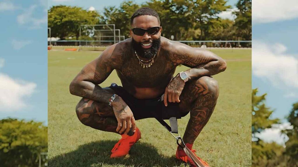Odell Beckham Jr in sunglasses and silver necklace squats on a sunny grassy field in red sneakers, grinning.