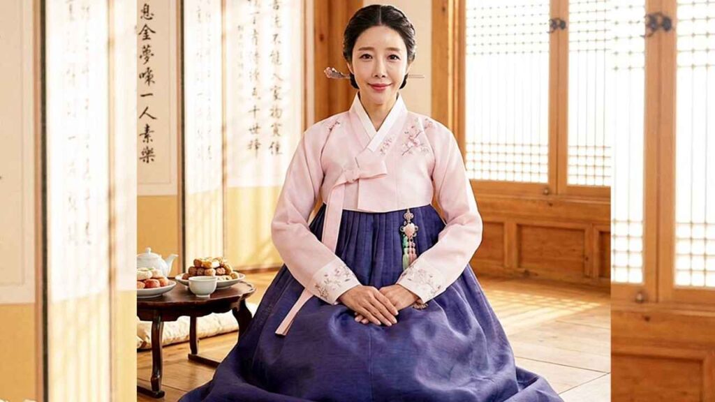 Oh Seung-eun in a pink jeogori and navy hanbok sits on a wooden floor in a sunlit traditional room, hands folded in her lap, with a tea set and pastries on a small table beside her and vertical calligraphy scrolls in the background.