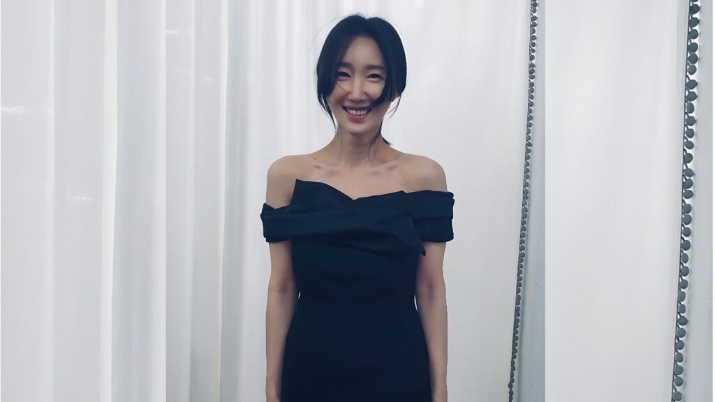 Oh Yeon-ah in an off-shoulder black dress poses before white vertical curtains, a simple elegant backdrop.