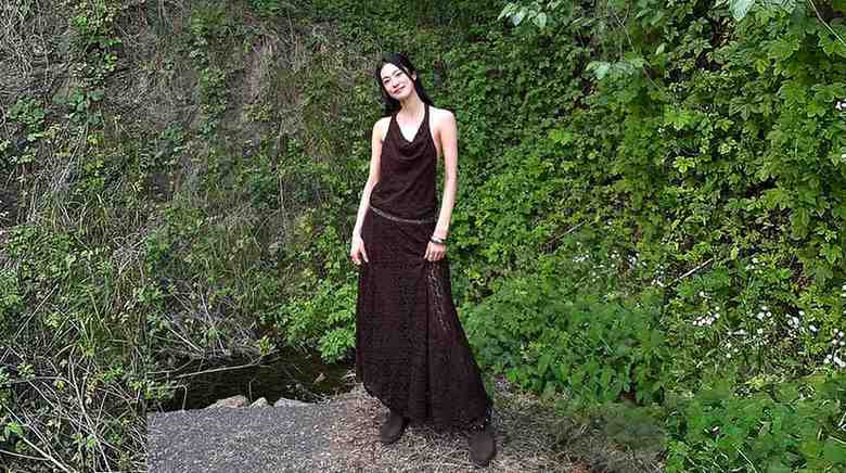 Ok Ja-yeon (옥자연) in a dark brown halter dress and brown boots stands on a stone path beside a lush ivy-covered wall.