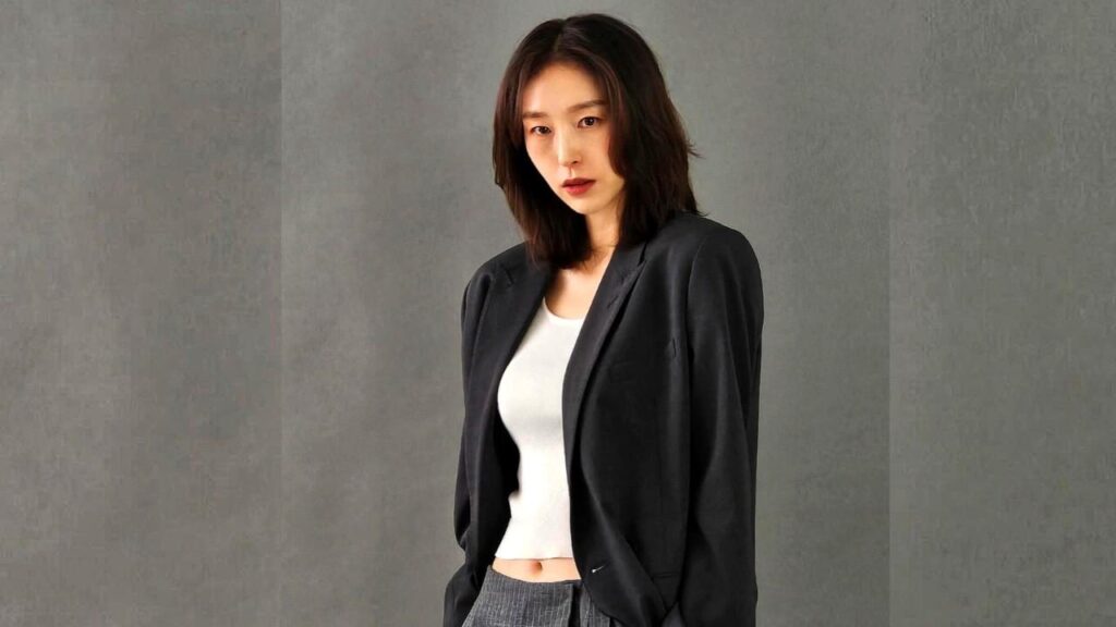 Ok Ji-young with shoulder-length dark hair wears a black blazer over a white crop top, gray trousers, against a textured gray wall.