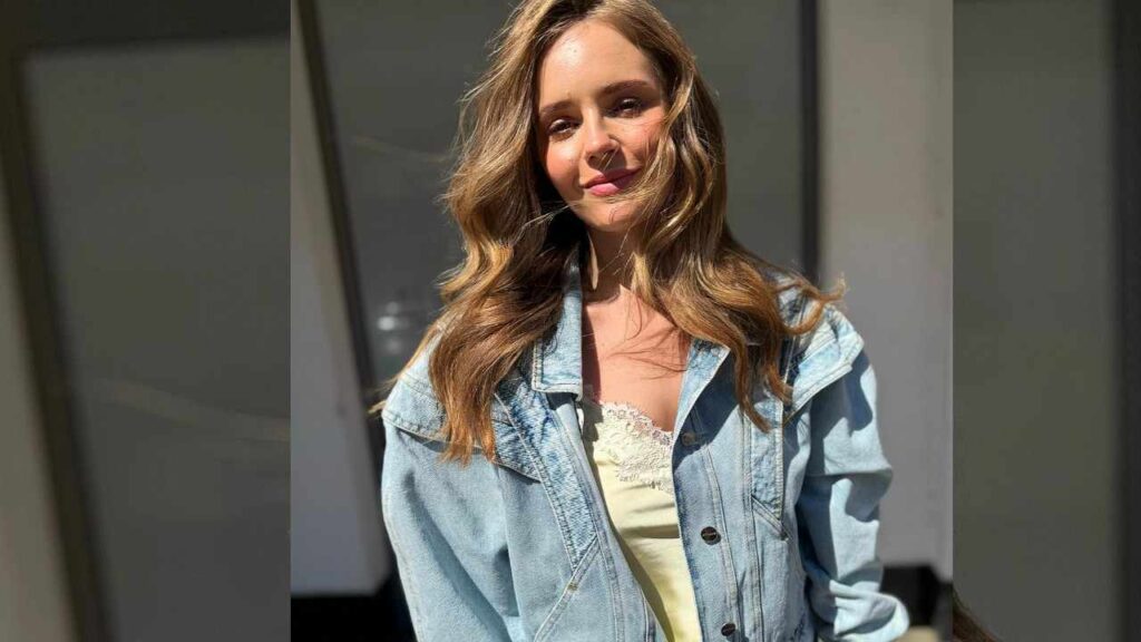 Olga Kalicka with wavy, light brown hair, wearing a cream lace top under a light denim jacket outdoors.
