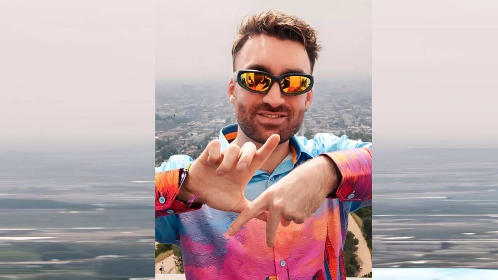 Oliver Heldens in a vibrant pink-to-blue gradient shirt and orange mirrored sunglasses gestures with his hands, cityscape below.