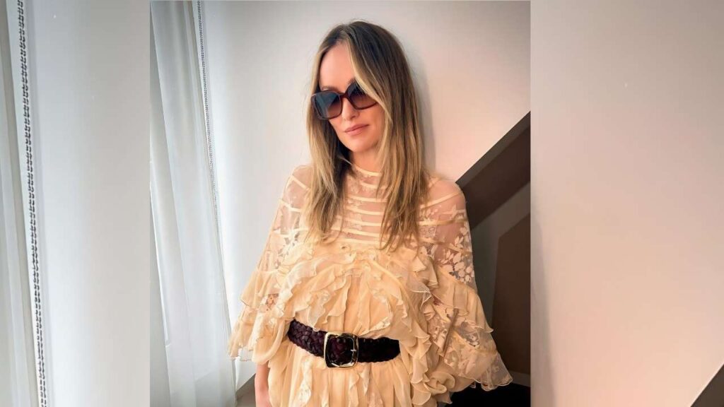 Olivia Wilde wearing sunglasses sports a sheer beige lace top with cascading ruffles and a wide belt, posing by a window.