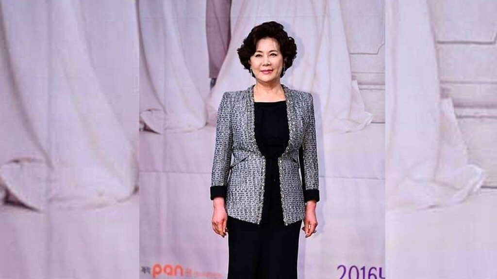 Park Jung-soo with short curly hair in a grey tweed blazer over black, standing before pale draped backdrop.