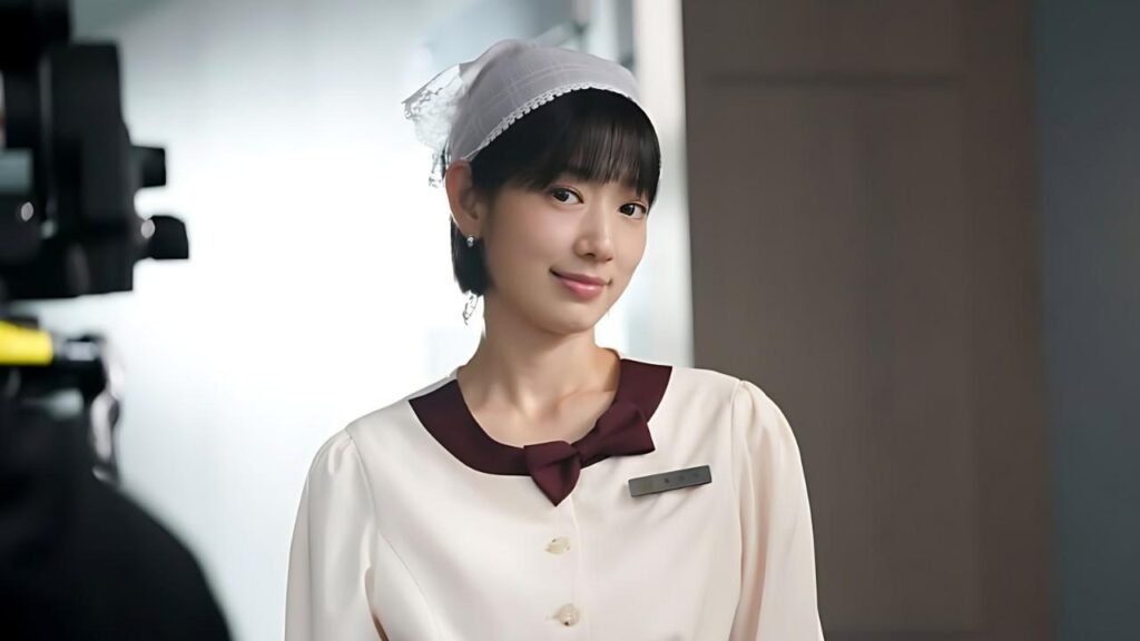 Park Shin-hye in a cream maid-style uniform with a white lace cap and burgundy bow at the collar, name tag, posing near a camera on a studio set.