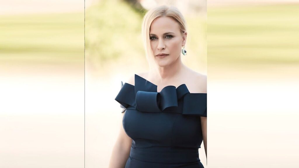 Patricia Arquette in a dark blue dress with a large bow at the neckline, standing outdoors with a soft background.