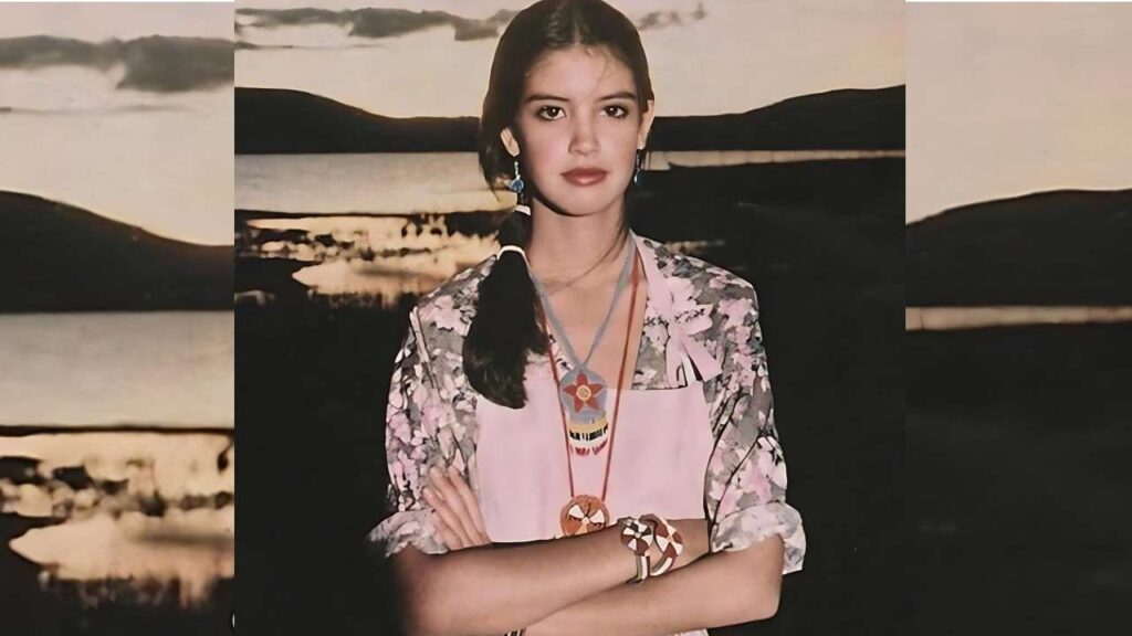 Phoebe Cates crosses arms, wearing an apron over a floral top and blue earrings, in front of sunset lakeside backdrop.