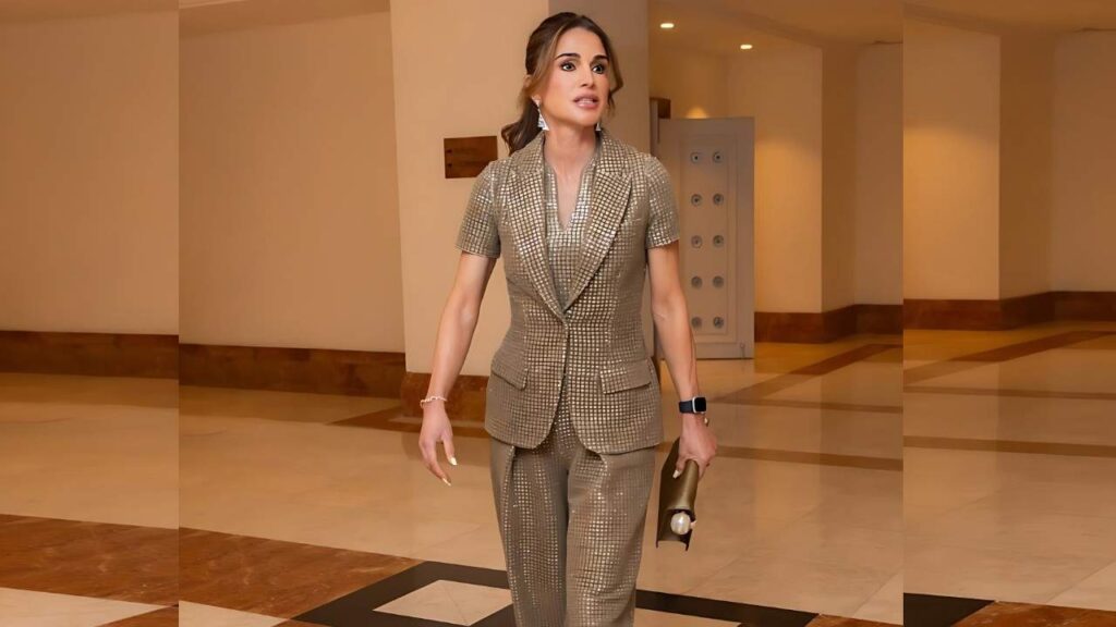 Queen Rania Al Abdullah in a tan checkered blazer and pantsuit walks through a warm marble lobby, holding a gold clutch.
