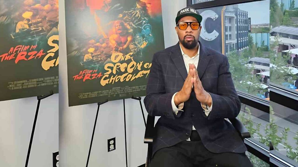 RZA in a dark checkered blazer and Park Hill cap with sunglasses sits with hands pressed in prayer, posters behind him.
