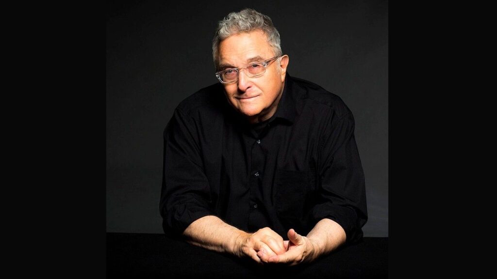 Randy Newman with gray hair and glasses in a black shirt, hands clasped on a dark table against a black backdrop.