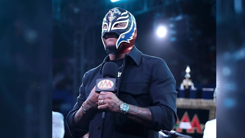 Rey Mysterio in blue-white-red mask speaks into a mic with AAA logo; dark shirt, tattooed arms, backdrop with a trophy.