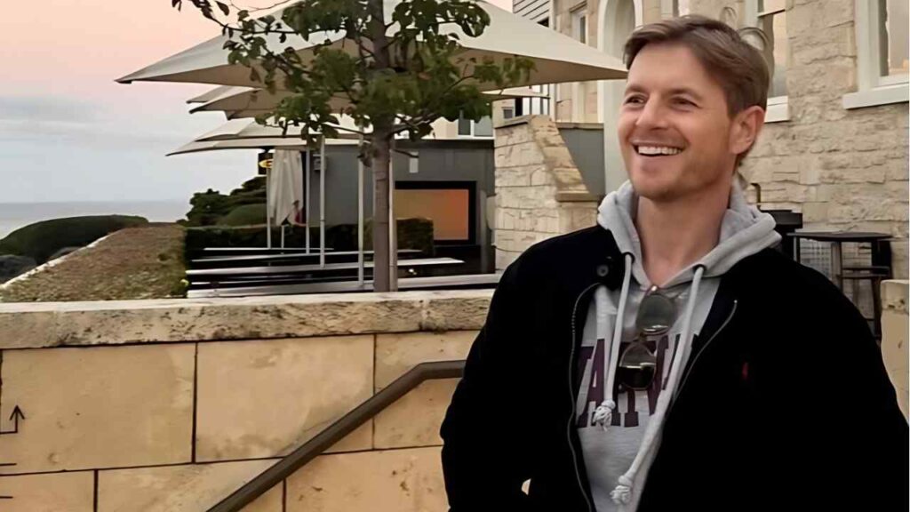 Rick Cosnett in gray hoodie and black jacket stands by a stone terrace, sunglasses clipped to his hoodie, with sea behind.