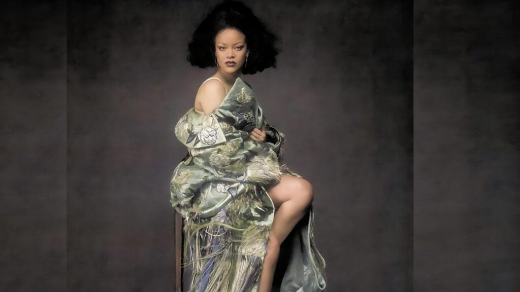 Rihanna with curly black hair wearing a pale green floral kimono, sitting with one leg up, dark studio backdrop.