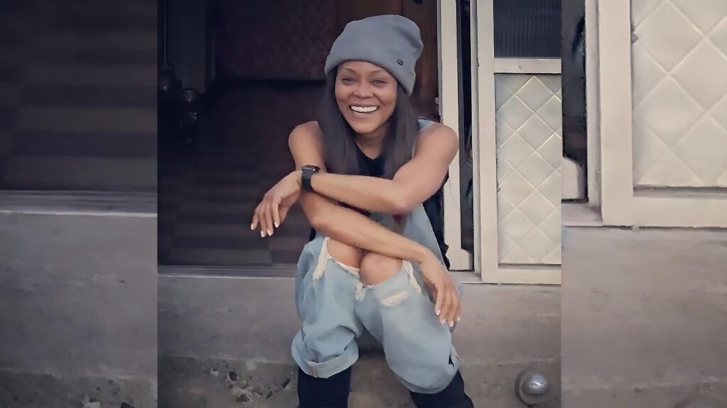 Robin Givens crouches on concrete steps outside a doorway, wearing a gray beanie, sleeveless top, ripped light-blue jeans, and a smartwatch.