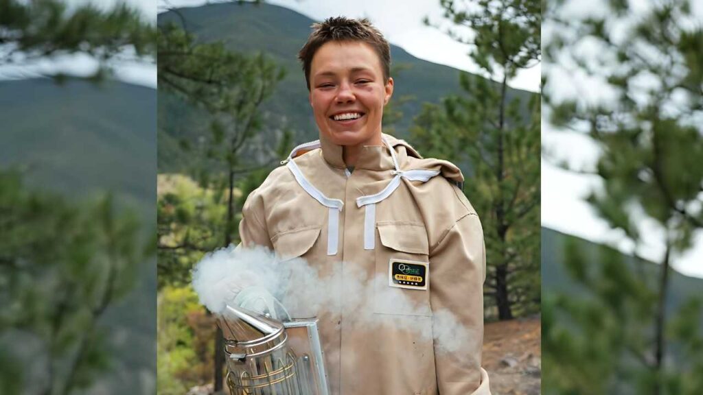 Rose Namajunas in beige protective suit with white trim outdoors, holding a metal device that releases smoke among pine trees.