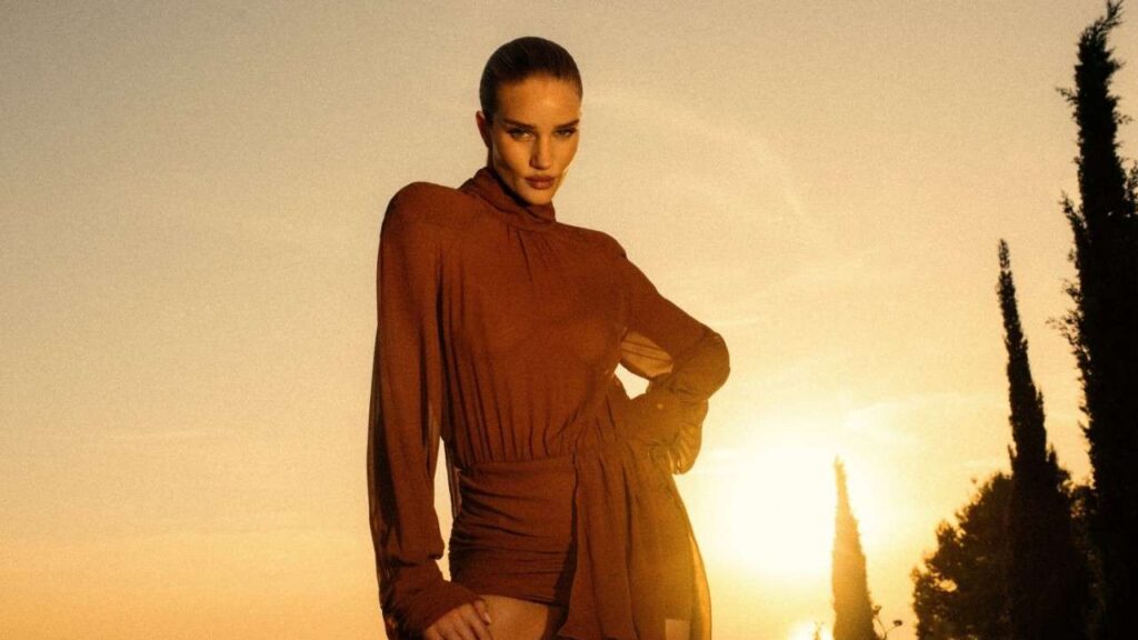 Rosie Huntington Whiteley in a rust belted blouse with billowy sleeves poses at sunset, hand on hip, warm outdoor scene with cypress trees