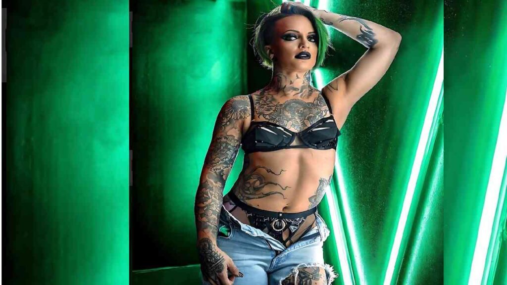 Ruby Riott with short green hair wearing a black bra and ripped jeans, striking a pose against neon green light tubes.