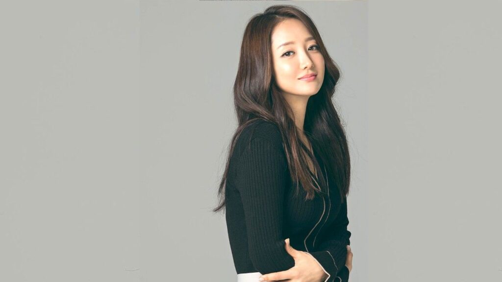 Sa Kang (사강) with long dark hair and a soft smile, wearing a black ribbed sweater, stands against a light gray backdrop.