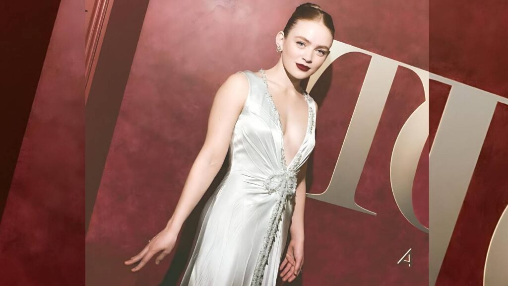 Sadie Sink in a plunging silver-white gown with a jeweled waist poses against a maroon backdrop with large gold letters.