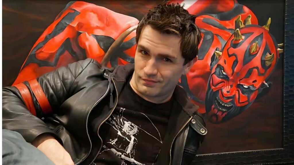 Sam Witwer with short dark hair wearing a black graphic tee and leather jacket, leaning beside a vivid red demon mural.