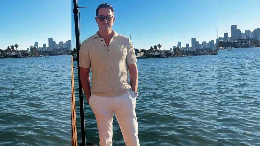 Sasha Roiz in beige polo and cream pants, stands by the water, hands in pockets, with a city skyline across the bay under blue sky.