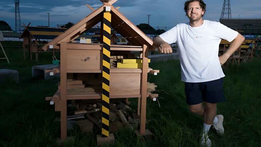 Sebastian Vettel in white t-shirt and shorts leans on a wooden miniature shed with a black-and-yellow striped post, outdoors on grass.