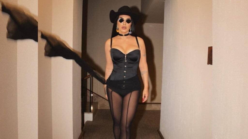 Seka Aleksić in a black corset, sheer tights, and garter belt walks down a beige hallway, wearing a black hat, sunglasses, and choker.