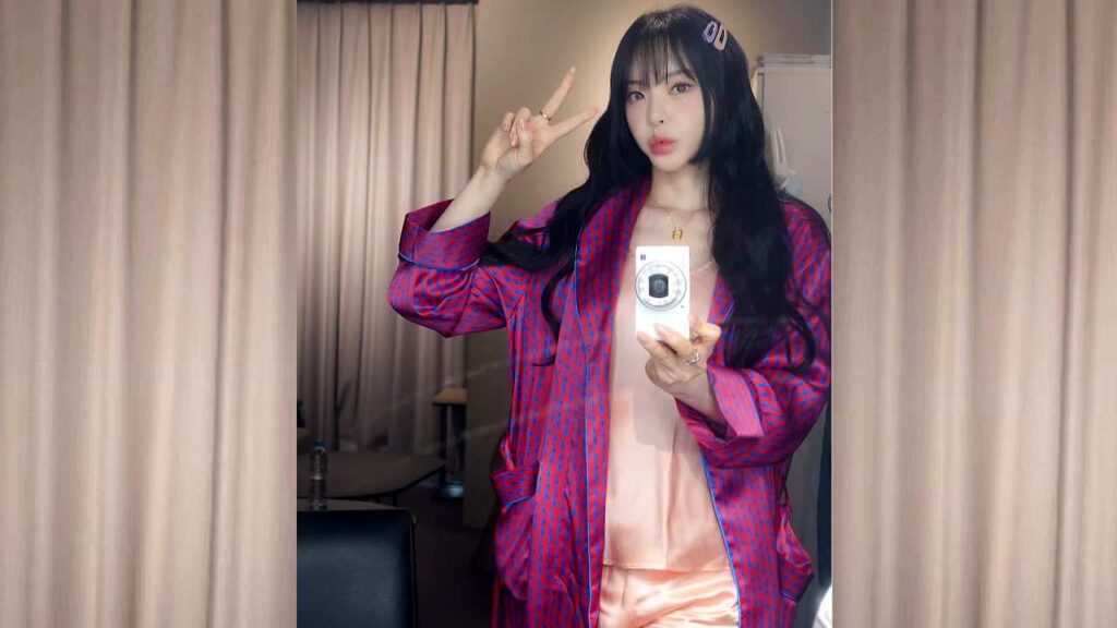 Seo Yu-ri poses in a mirror, flashing a peace sign while holding a white camera, wearing pink satin and a purple robe.