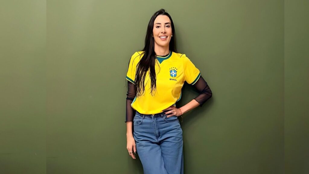 Sheilla Castro stands against an olive-green wall in a yellow Brazil soccer jersey with sheer black sleeves, blue jeans, hand on hip.