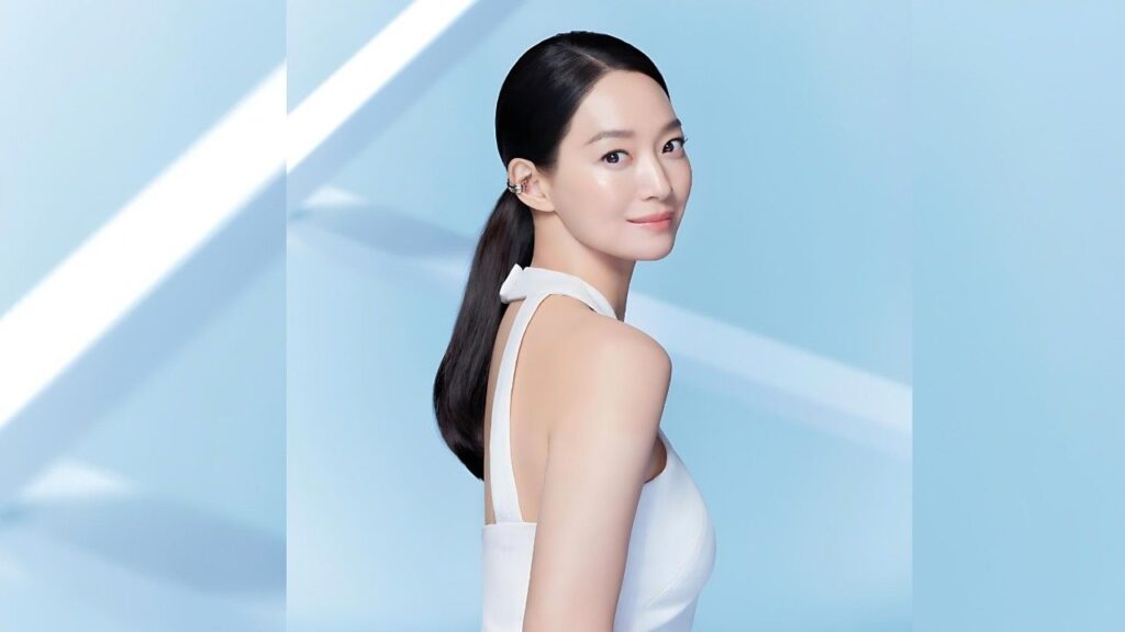 Shin Min-a with sleek dark hair in a white halter dress, posed against a blue studio backdrop with light stripes.