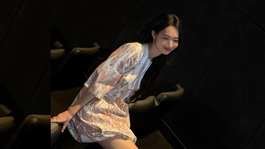 Shin Min-a with long black hair wearing a pale pastel floral dress sits against a dark geometric wall, smiling softly.