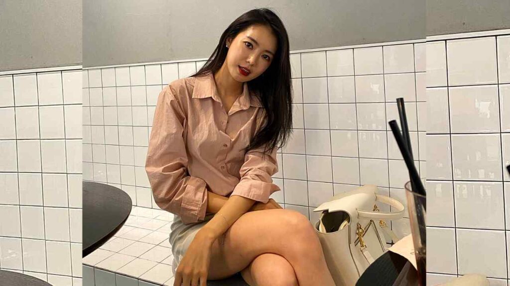 Shin Min-hee with long dark hair sits cross-legged in a peach blouse; a white handbag rests beside her.