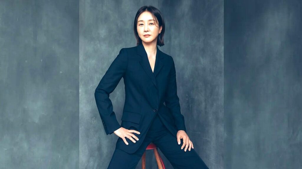 Shin So-mi in a tailored navy suit sits on a wooden stool against a textured blue-gray backdrop, hands poised confidently.